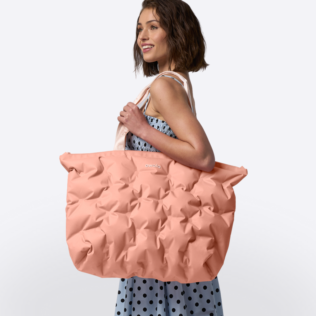 Model carrying the Owala Cloud tote in Rosewood on her shoulder, coral pink puffer-quilted body with blush owala-tape handles